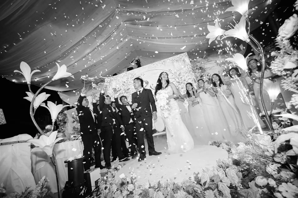 Black-and-white photo of the couple and their entourage on a flower-filled stage as confetti falls