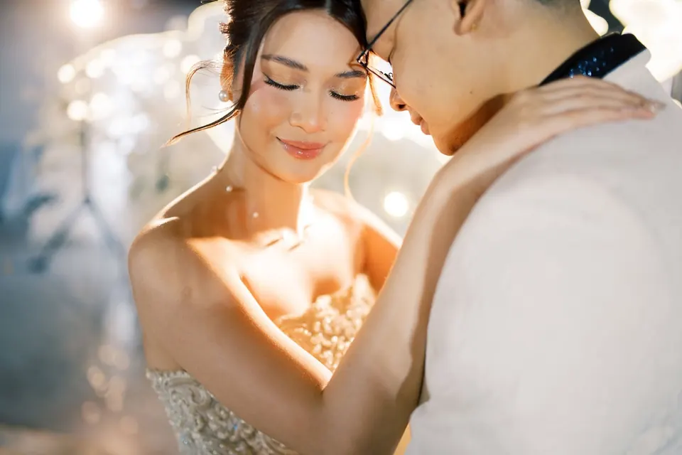 The bride smiles with her eyes closed as the groom holds her close in warm golden light